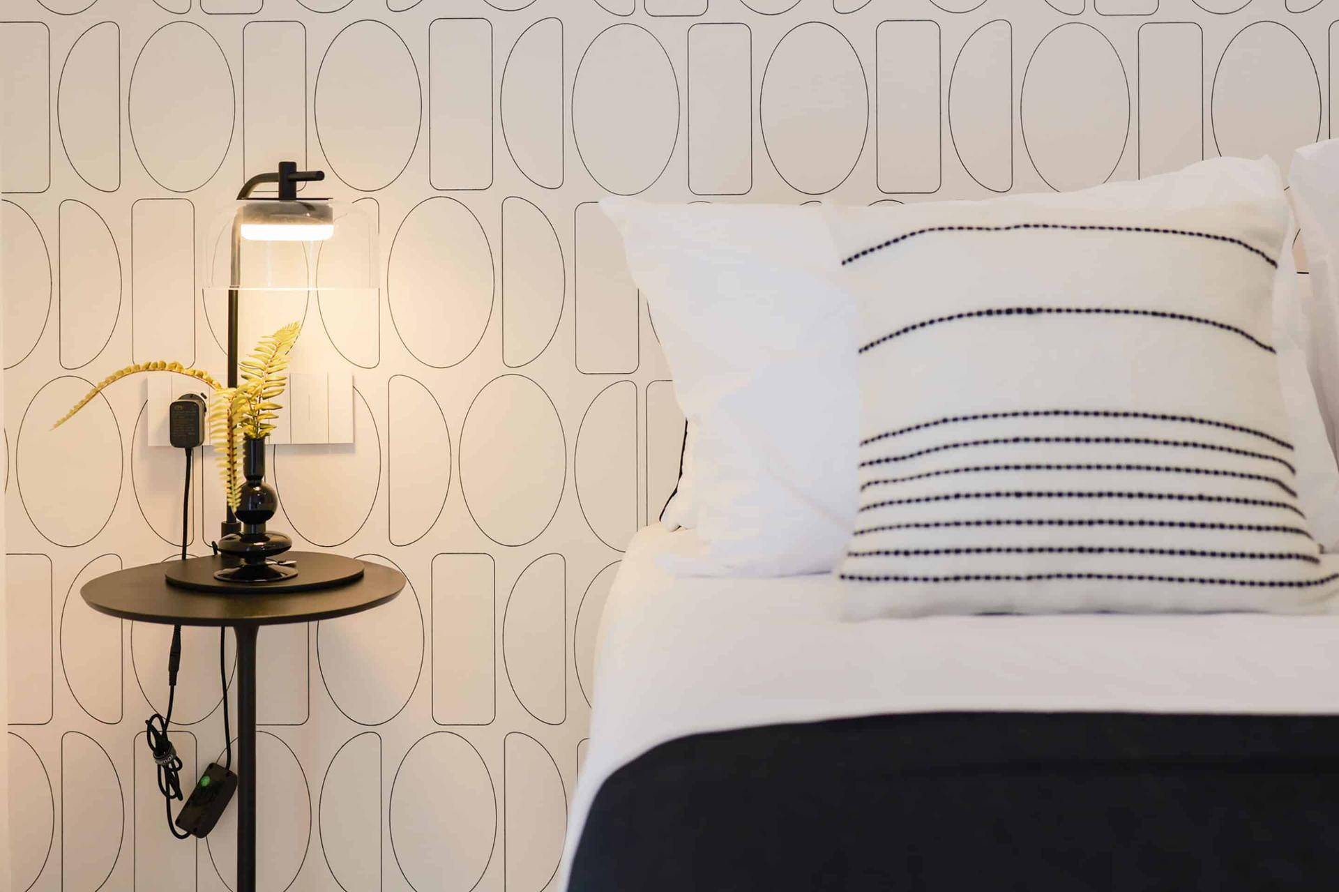 Schlafzimmerdetail mit modernem Nachttisch, Lampe und grafischer Tapete.