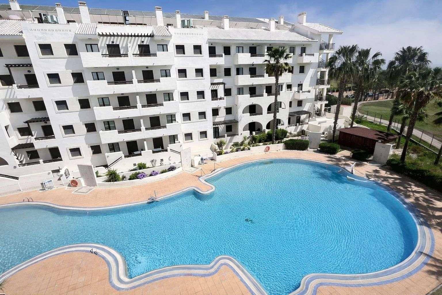 Luftaufnahme eines weißen Apartmentkomplexes mit großem, geschwungenem Swimmingpool und Palmen.