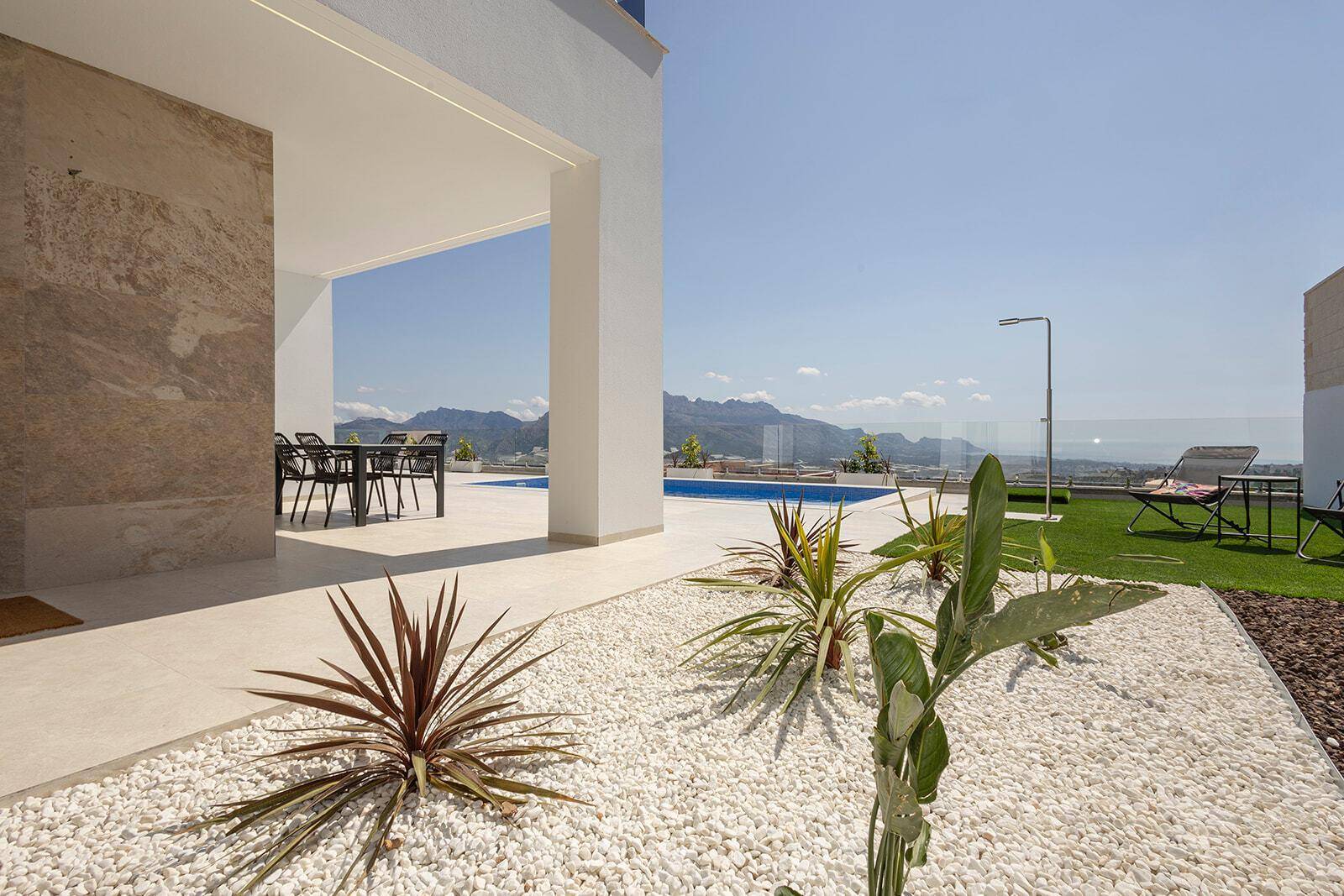 Gartenbereich mit Kies, Pflanzen, Kunstrasen und Blick auf den Pool und die Berge.