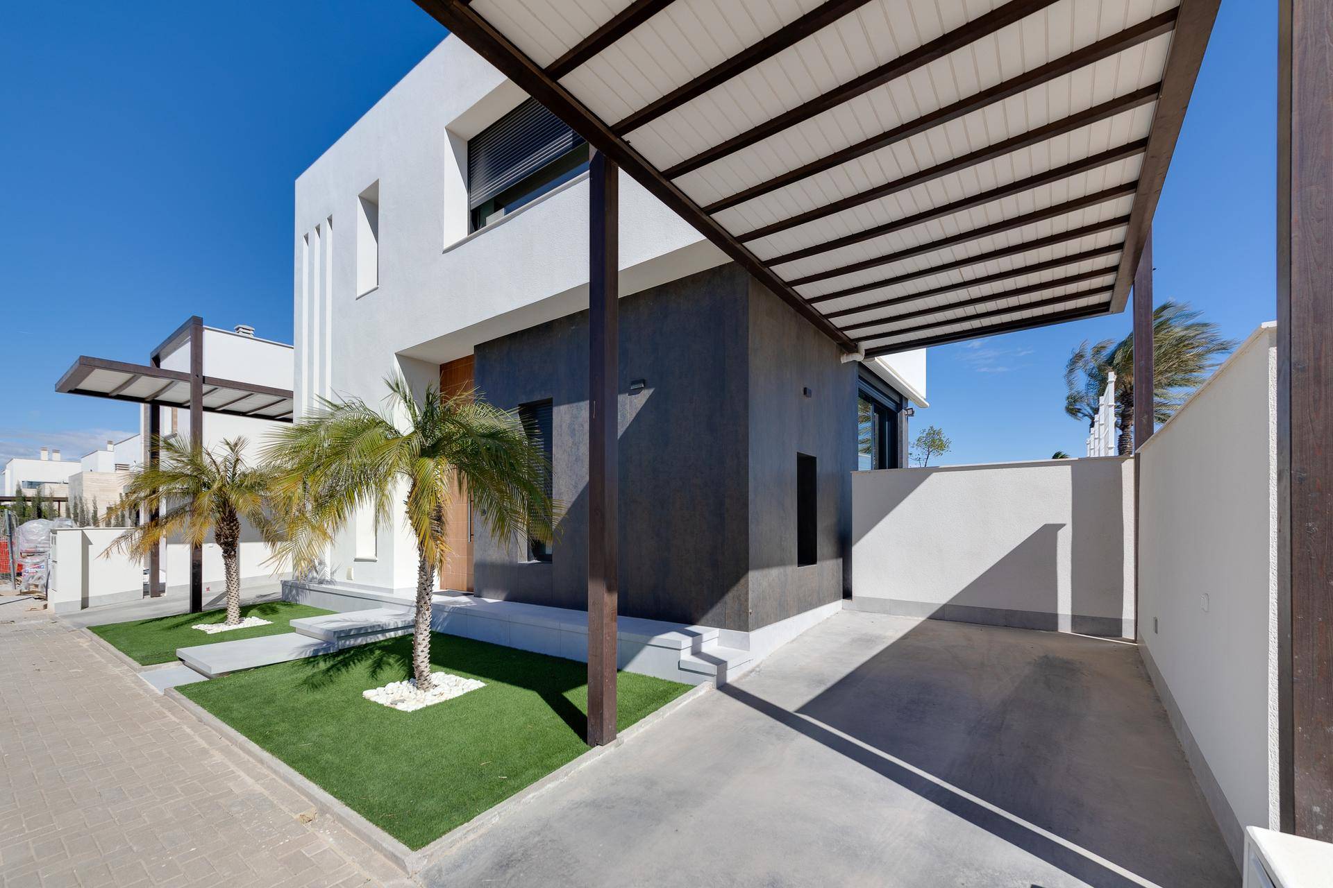 Moderne Hausfassade mit klaren Linien, Holztür und überdachtem Parkplatz unter blauem Himmel.