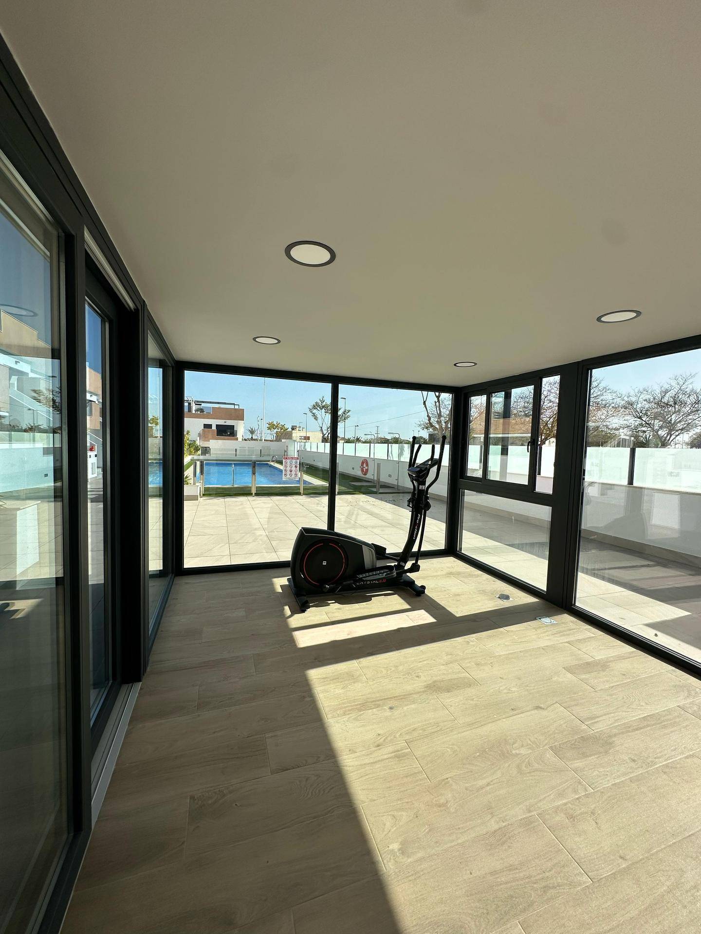 Heller Fitnessraum mit Ellipsentrainer und großen Fenstern mit Blick auf den Poolbereich.