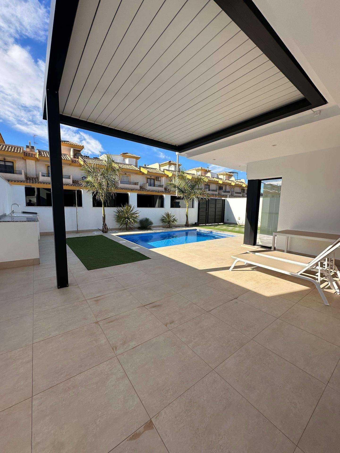 Überdachte Terrasse mit Liegestuhl und Blick auf den Pool und die umliegenden Häuser.