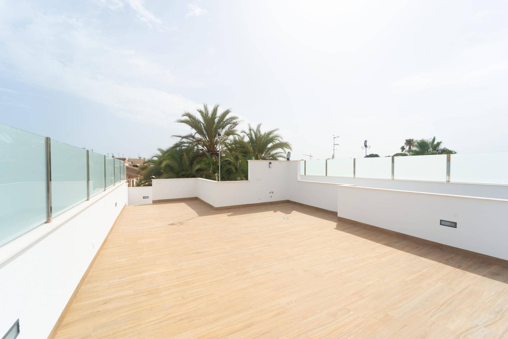 Weitläufige Dachterrasse mit hellen Fliesen und Glasbrüstung, umgeben von Palmen unter blauem Himmel.