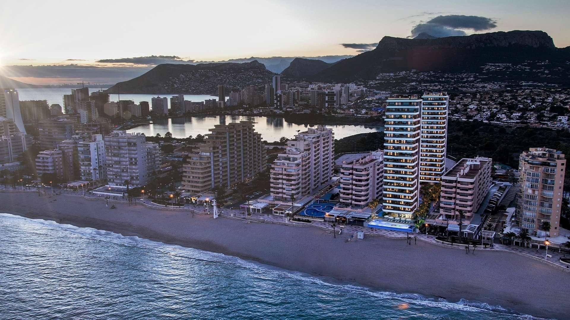 Abendliche Luftaufnahme der Küstenstadt mit beleuchteten Hochhäusern, Strand und malerischem Sonnenuntergang über den Bergen.