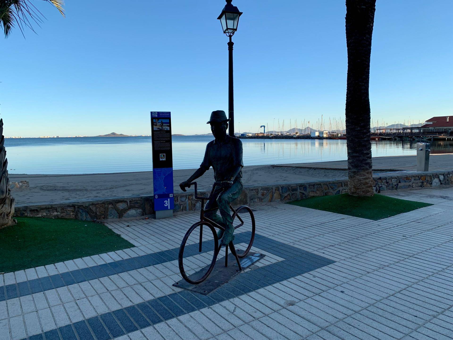 Statue eines Radfahrers auf einer Promenade mit Meerblick und Palmen im Hintergrund.