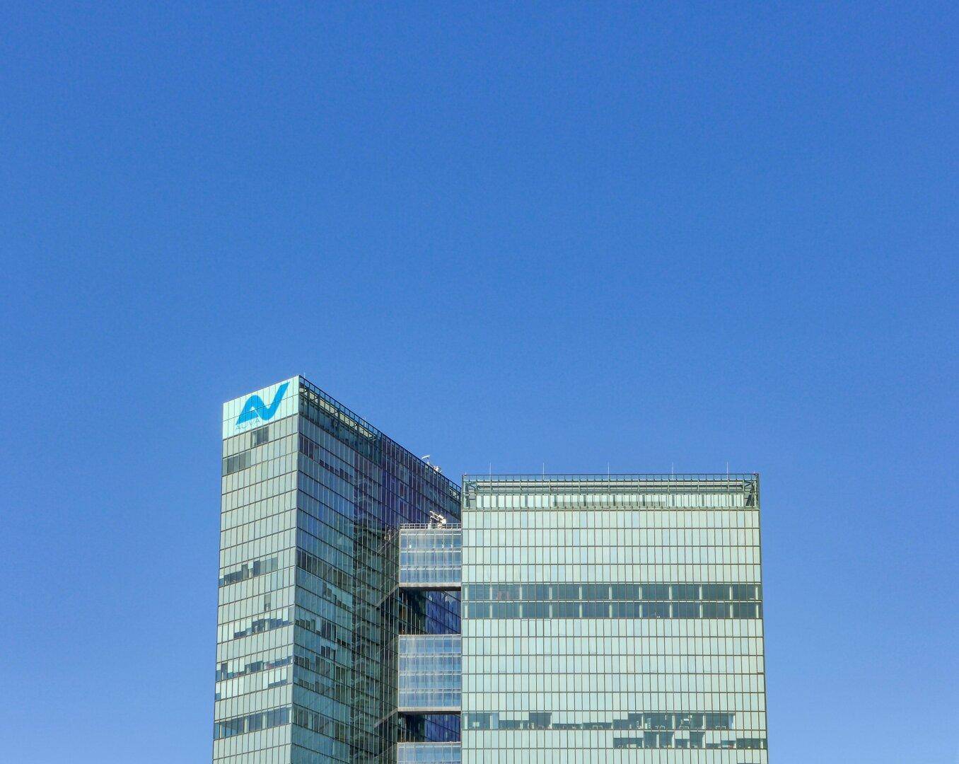 Oberer Teil eines modernen Hochhauses mit Glasfassade und Firmenlogo vor klarem blauem Himmel.