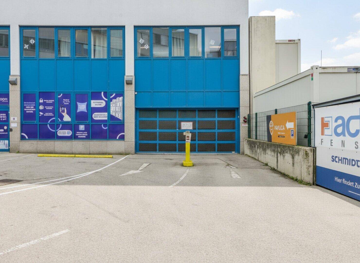 Erdgeschoss eines Bürogebäudes mit blauer Fassade, Garagentor und Parkplatz.