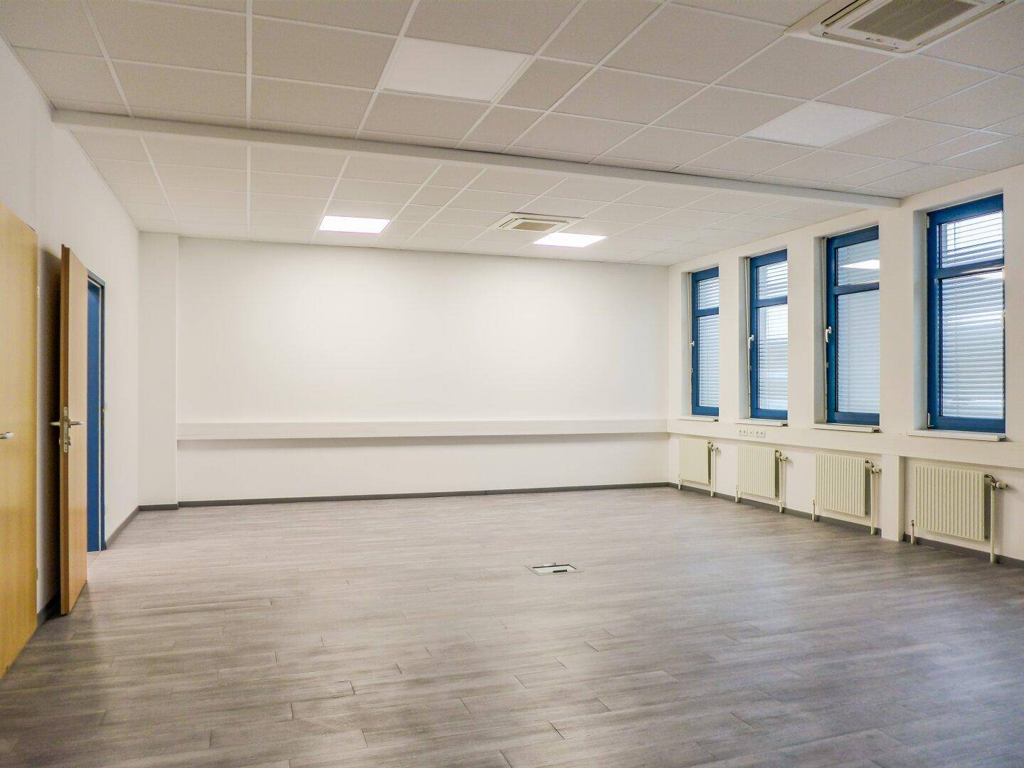Geräumiger, leerer Büroraum mit großen Fenstern, Heizkörpern und modernem Fliesenboden.