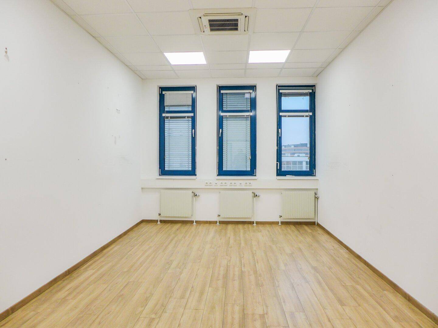 Heller, leerer Büroraum mit drei Fenstern, Heizkörpern und einem Boden in Holzoptik.