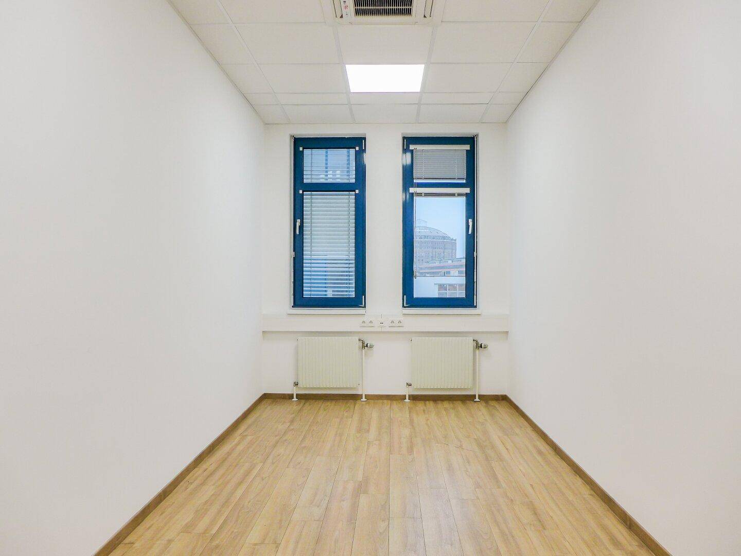Kleiner, heller Büroraum mit zwei Fenstern, Heizkörpern und einem Boden in Holzoptik.