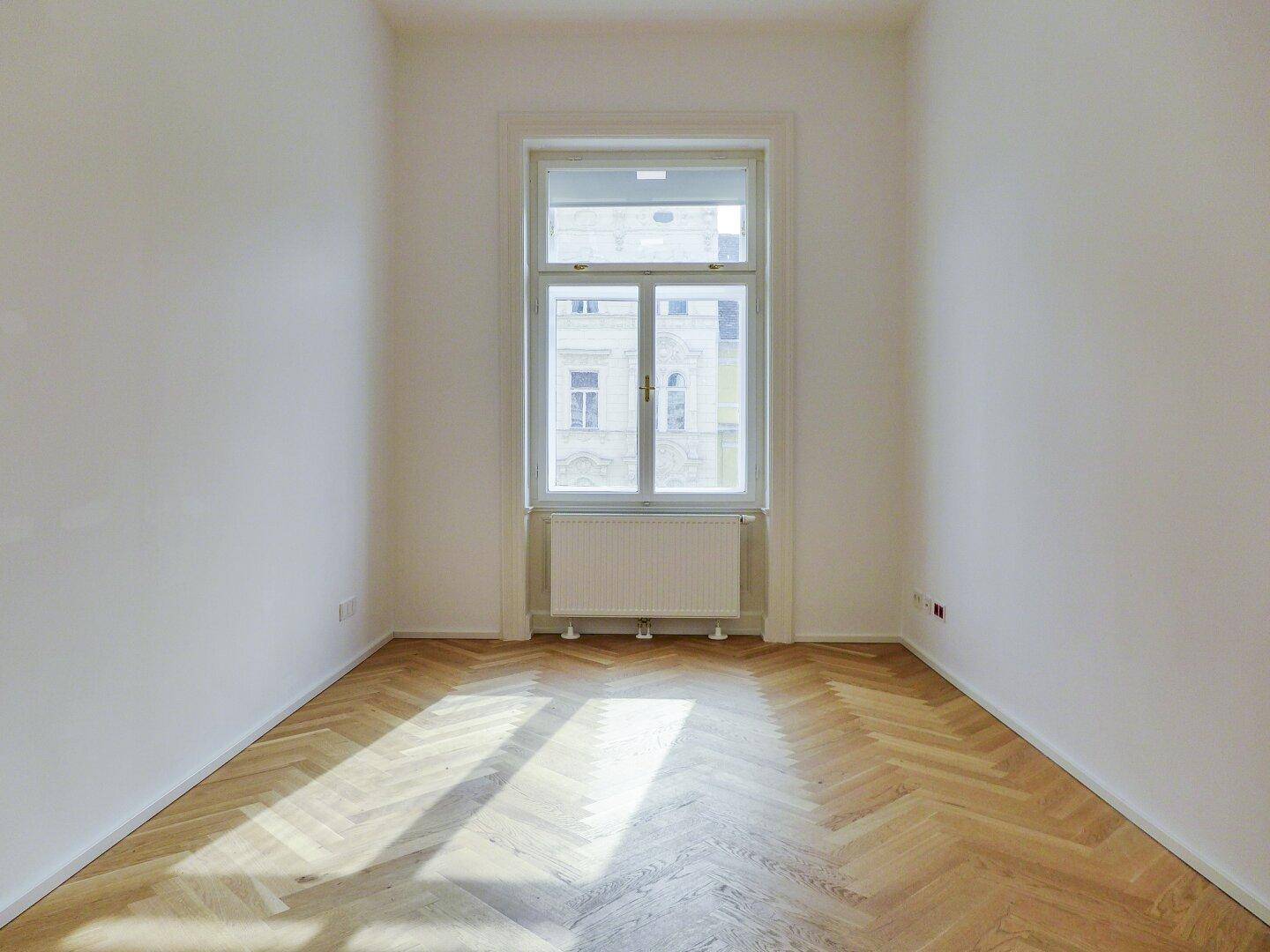 Schmaler, heller Raum mit einem großen Fenster und Fischgrätparkett, ideal als Arbeits- oder Schlafzimmer.