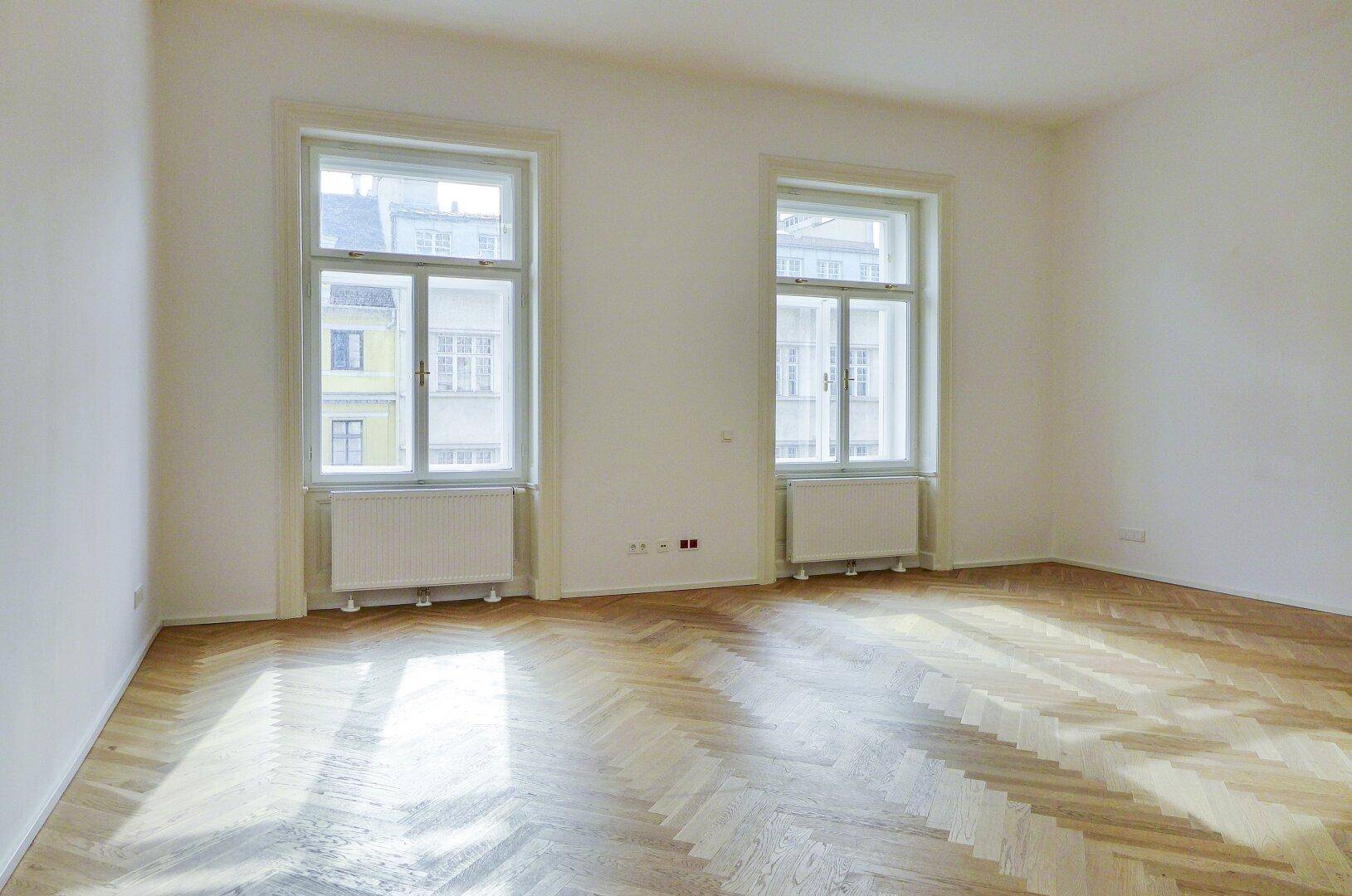 Geräumiger, heller Raum mit zwei großen Fenstern und elegantem Fischgrätparkett, ideal für vielfältige Nutzung.
