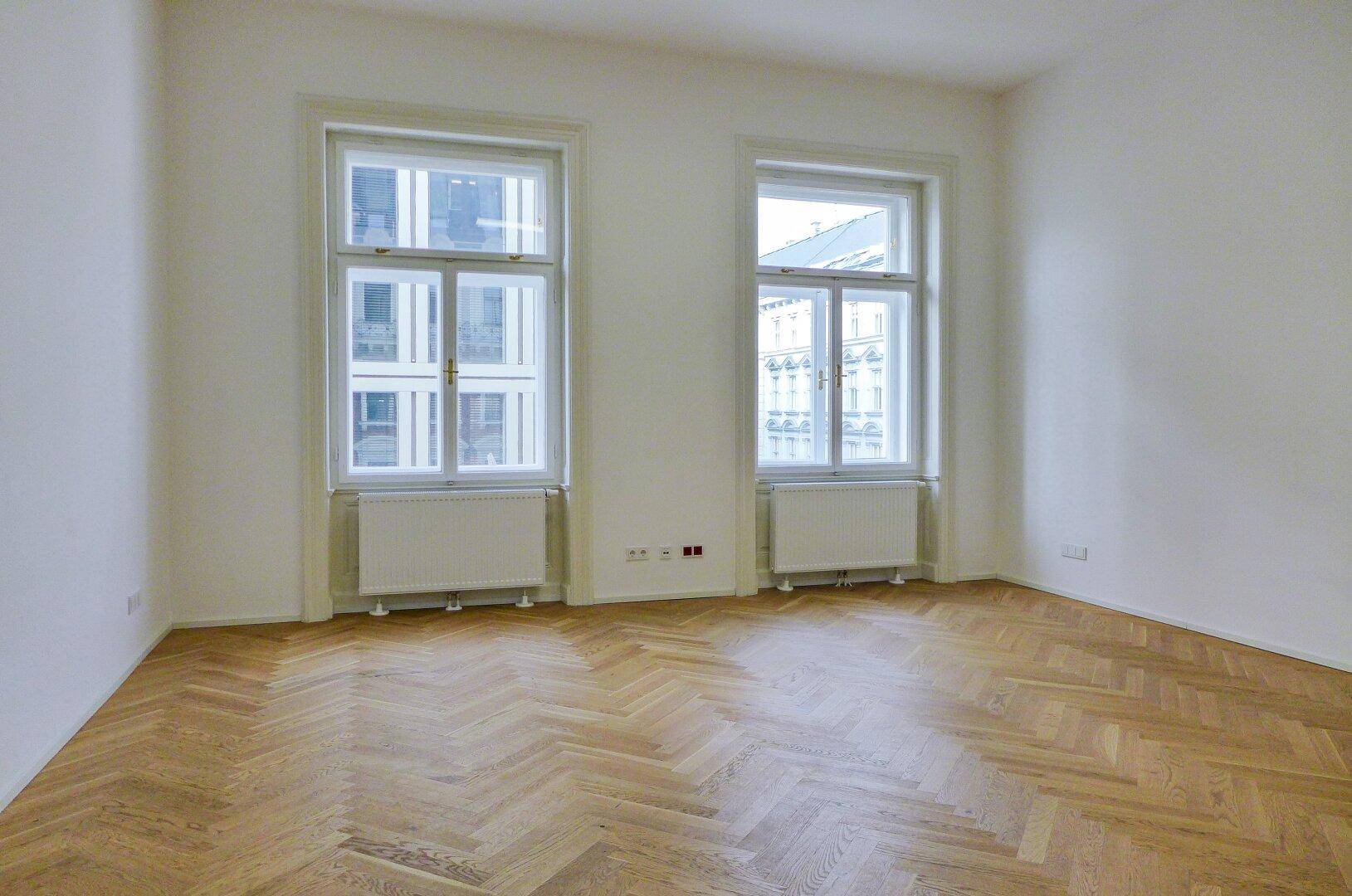 Heller Raum mit zwei hohen Fenstern und klassischem Fischgrätparkett, das viel Tageslicht reflektiert.