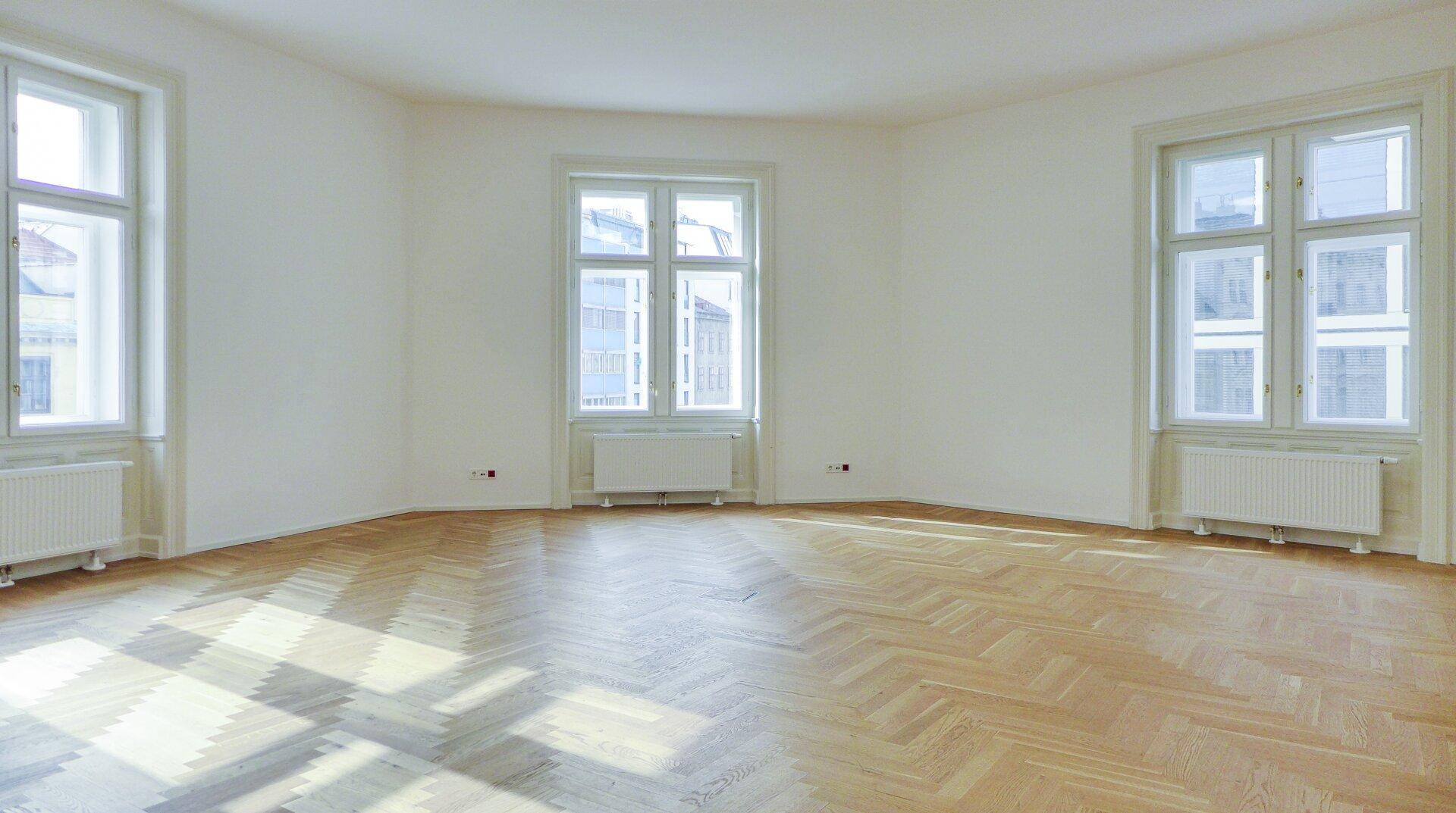 Lichtdurchfluteter Raum mit drei großen Fenstern und elegantem Fischgrätparkett, ideal für Wohnzwecke.