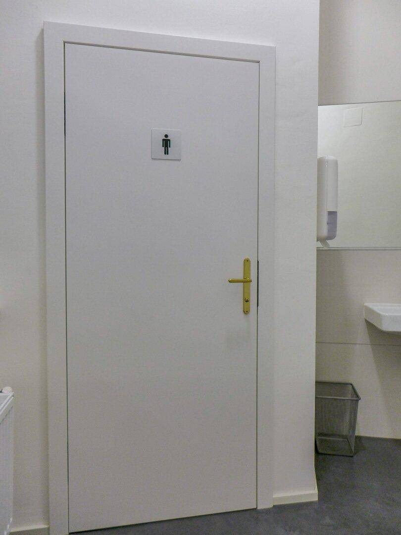 Eingang zu einem WC mit weißer Tür und Piktogramm, daneben ein Waschbeckenbereich mit Spiegel.