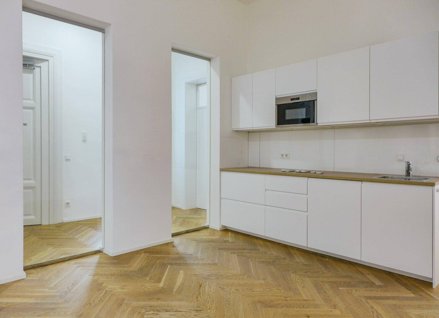 Moderne Einbauküche mit weißen Schränken, Holzarbeitsplatte und integrierter Mikrowelle auf Fischgrätparkett.