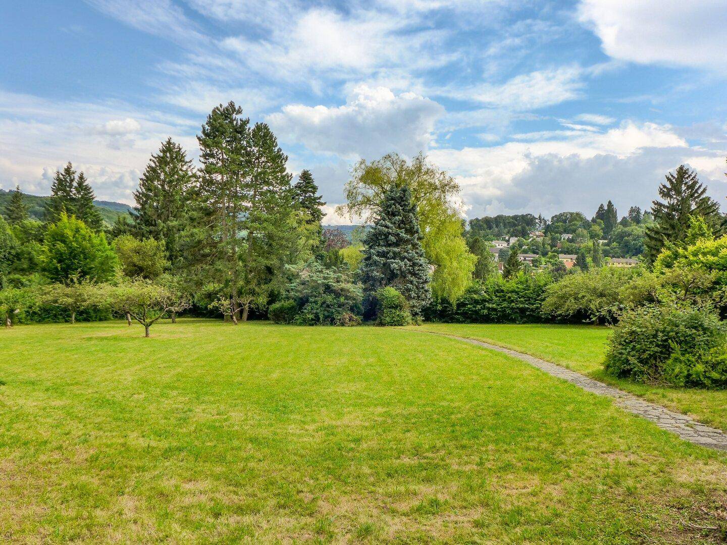 Großzügige Rasenfläche mit altem Baumbestand und einem geschwungenen Steinweg unter weitem Himmel.