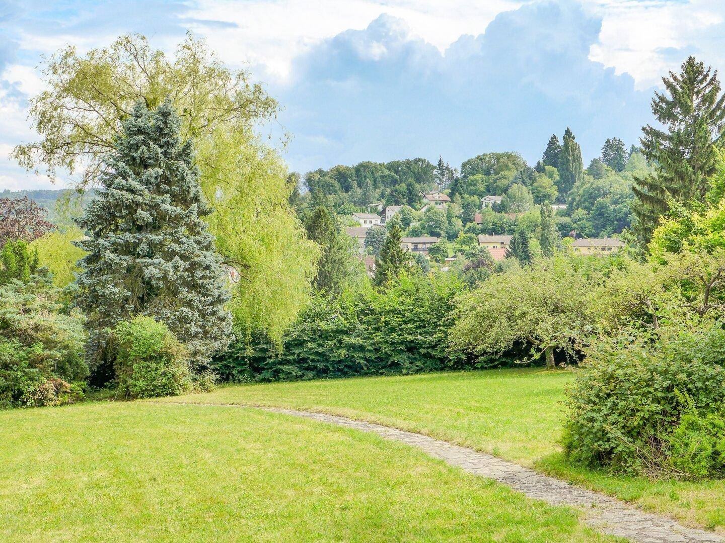 Blick über eine grüne Wiese und einen Steinweg auf bewaldete Hügel mit ländlicher Bebauung.