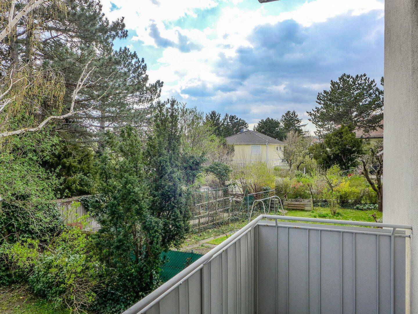 Blick vom Balkon ins Grüne mit Bäumen und einem Garten im Hintergrund, bietet eine ruhige Aussicht.