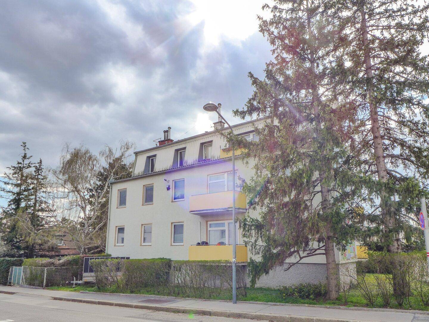 Mehrfamilienhaus mit gelben Balkonen und Bäumen im Vordergrund, an einer Straße gelegen.
