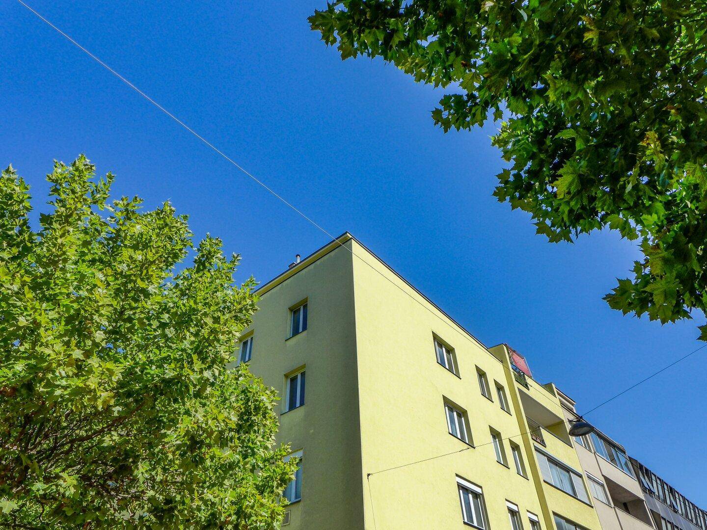 Gelbe Gebäudefassade mit mehreren Fenstern und Balkonen unter blauem Himmel und grünen Bäumen.