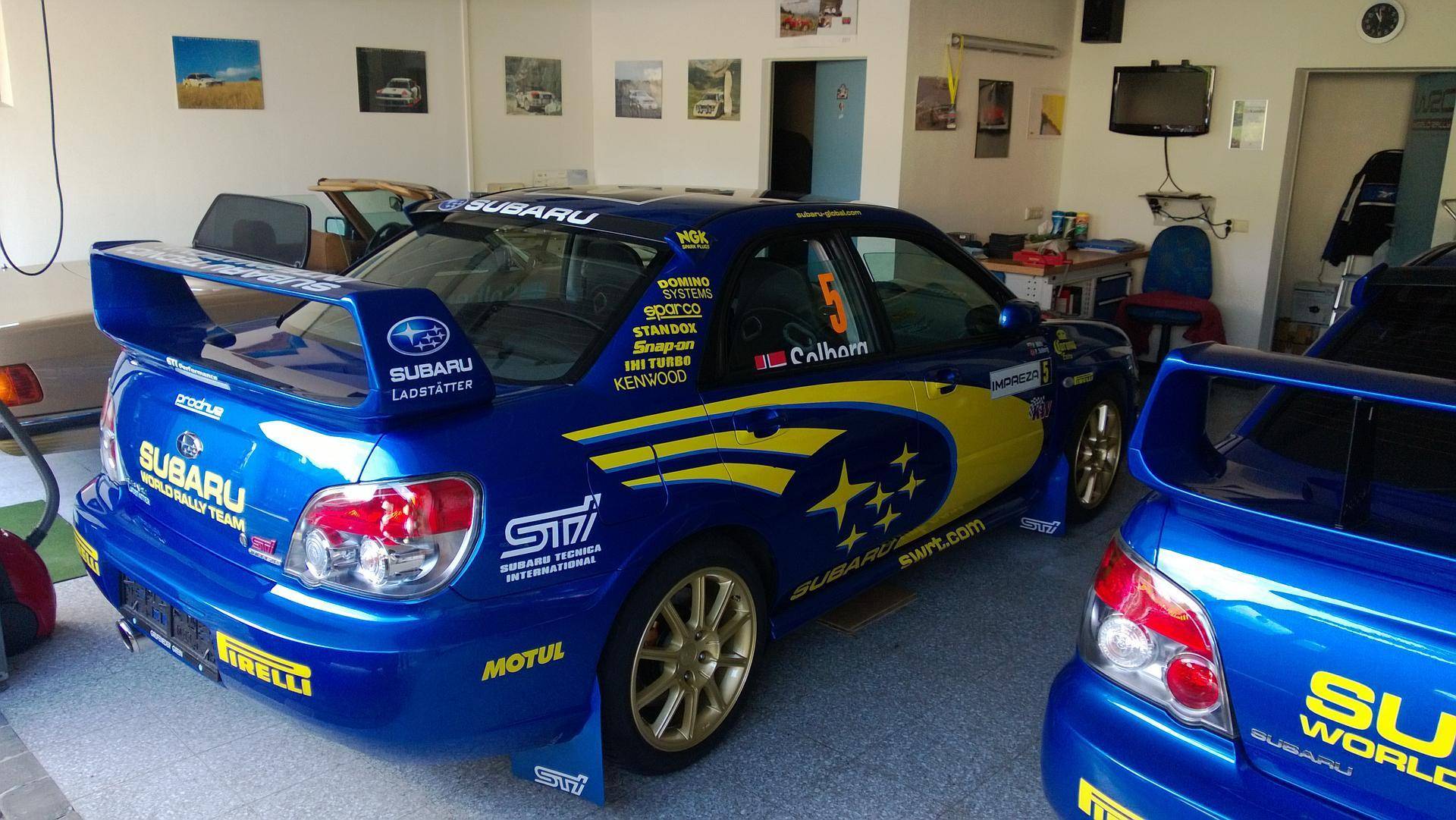 Geräumige Garage mit zwei blauen Rallye-Autos und Werkstattausrüstung an den Wänden.