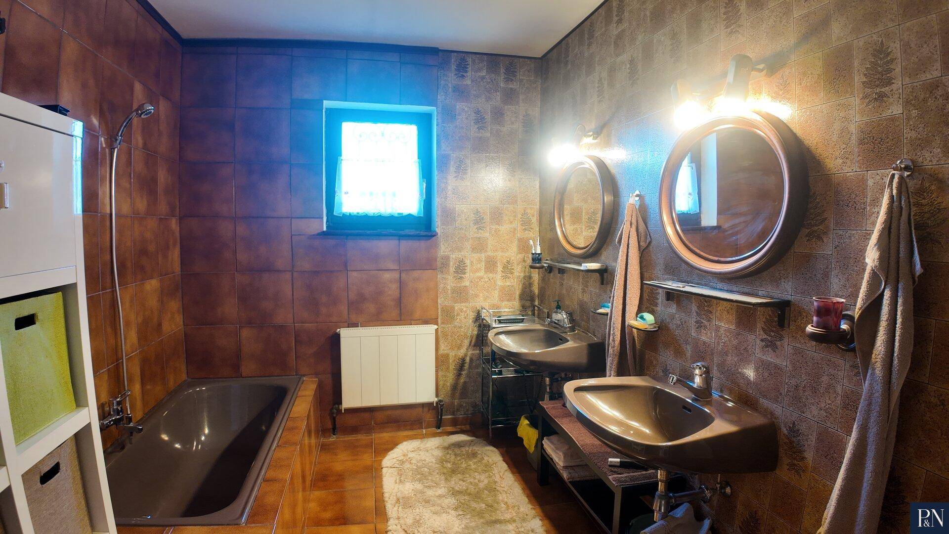 Badezimmer im 80er-Jahre-Stil mit braunen Fliesen, Badewanne und zwei Waschbecken.