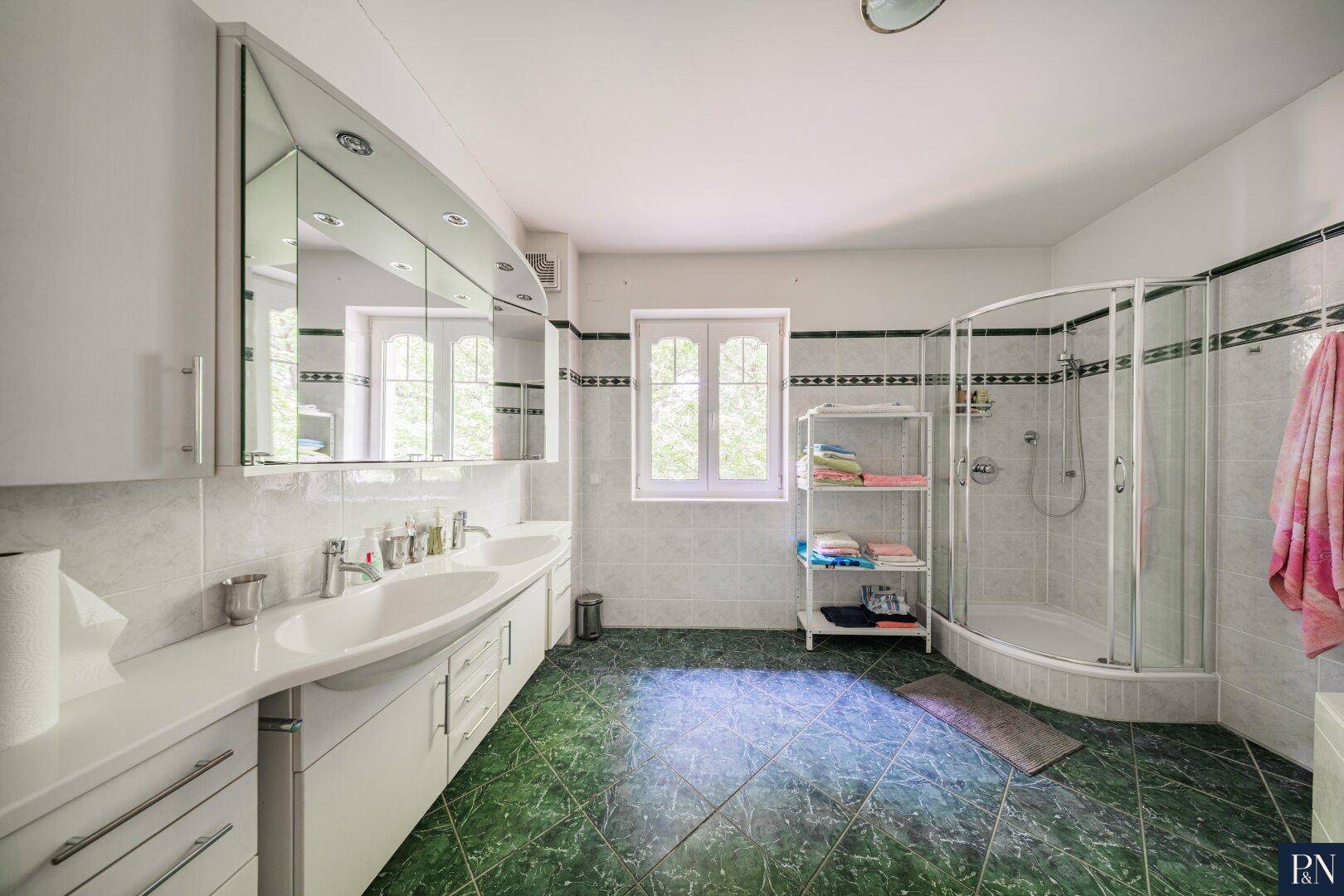 Helles Badezimmer mit Doppelwaschbecken, Duschkabine und Fenster mit Blick ins Grüne.