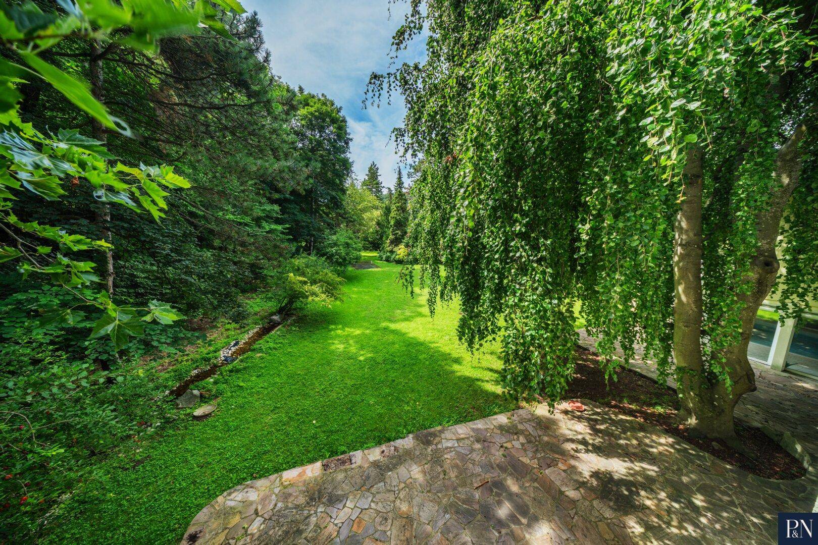 Panoramablick von der Terrasse auf den weitläufigen Garten und den Bachlauf.