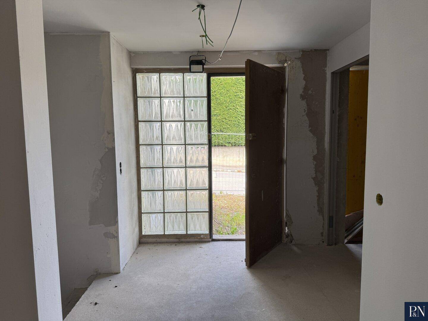 Eingangsbereich im Rohbau mit Glasbausteinen und offener Tür, die ins Freie führt.