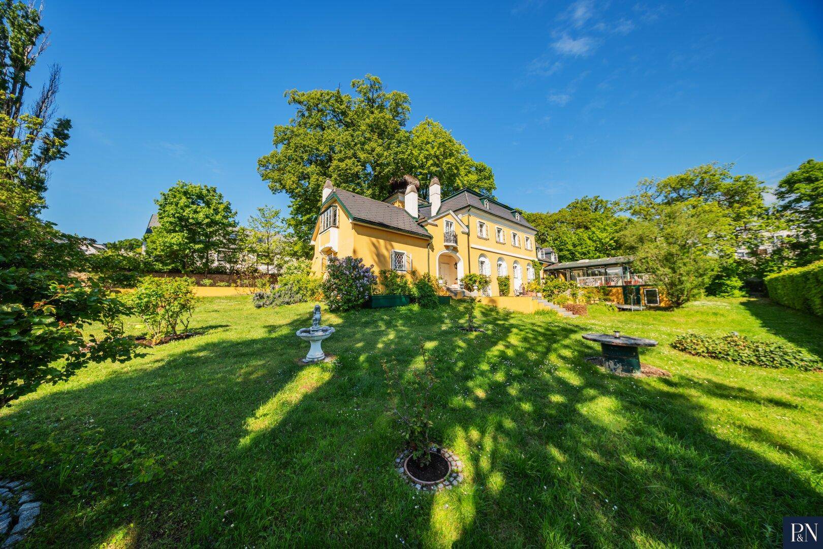 Großer, sonniger Garten mit Rasenflächen, Bäumen und einer dekorativen Gartenstatue.