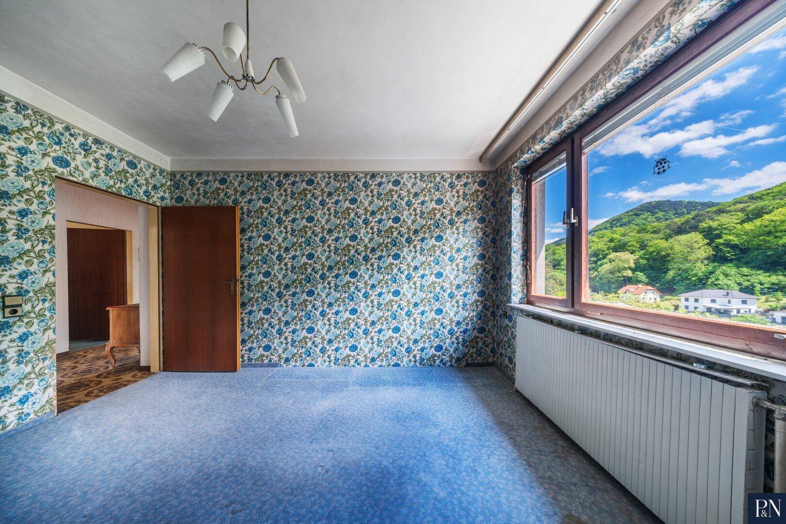 Zimmer mit auffälliger floraler Tapete, blauem Teppichboden und großem Fenster mit Bergblick.