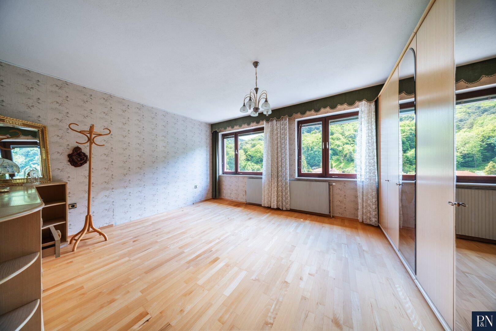 Helles Schlafzimmer mit Holzfußboden, Spiegelschrank und Blick auf die grüne Umgebung.