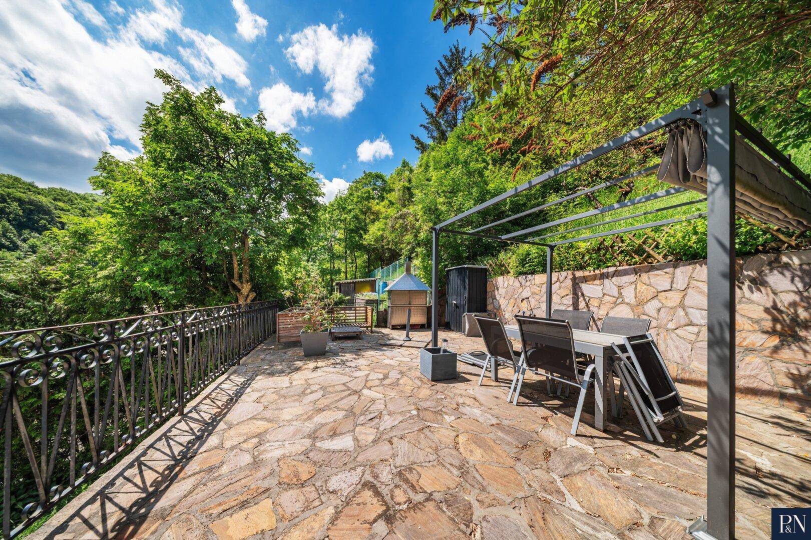 Große Steinterrasse mit Pergola, Gartenmöbeln und Blick auf die grüne Umgebung.