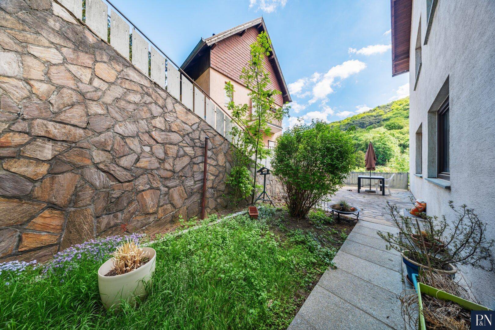 Kleiner Gartenbereich neben dem Haus mit Steinmauer und weiterer Terrasse im Hintergrund.