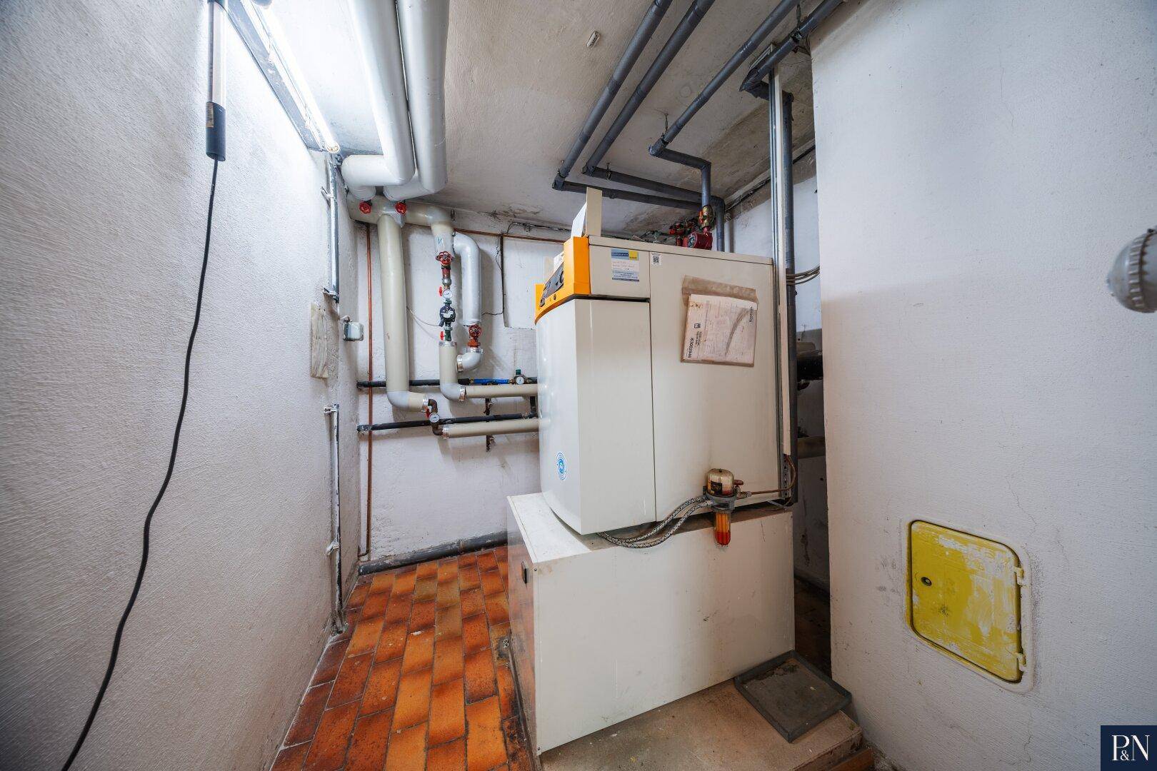 Heizungsraum mit Kessel, Rohren und Fliesenboden im Kellerbereich des Hauses.