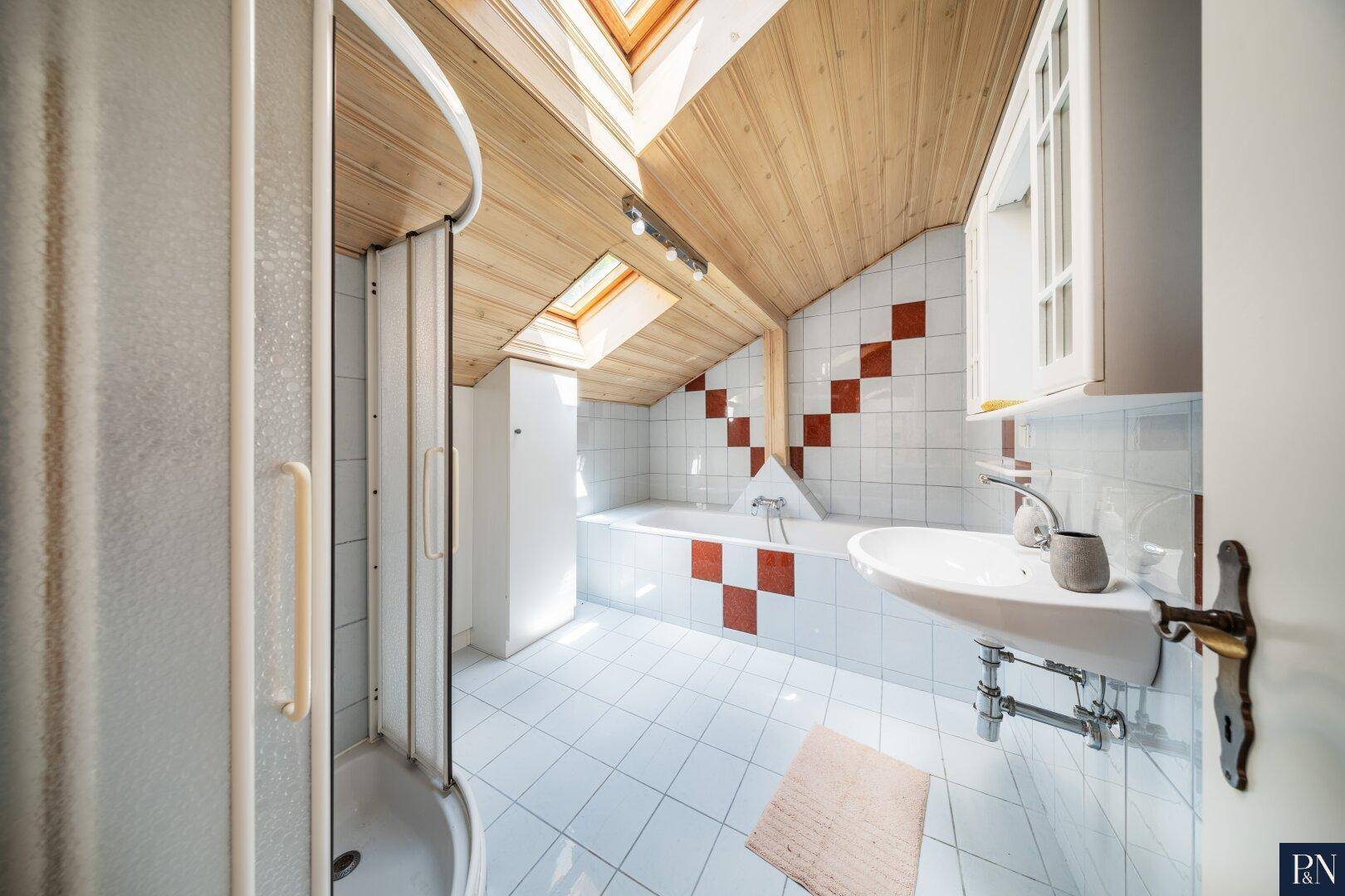 Badezimmer im Dachgeschoss mit Badewanne, Dusche, Dachfenstern und gemusterten Fliesen.