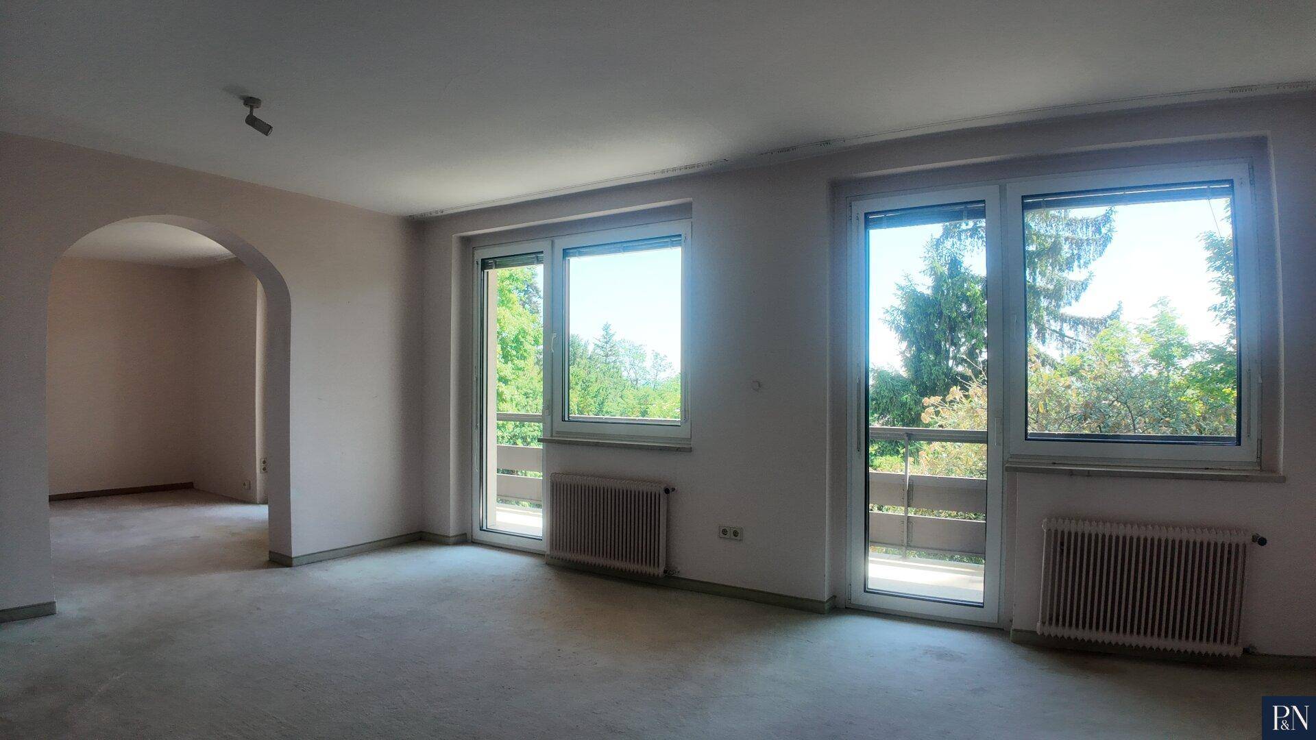 Heller Raum mit Bogenöffnung zu einem weiteren Bereich und zwei großen Fenstern mit Balkonzugang.