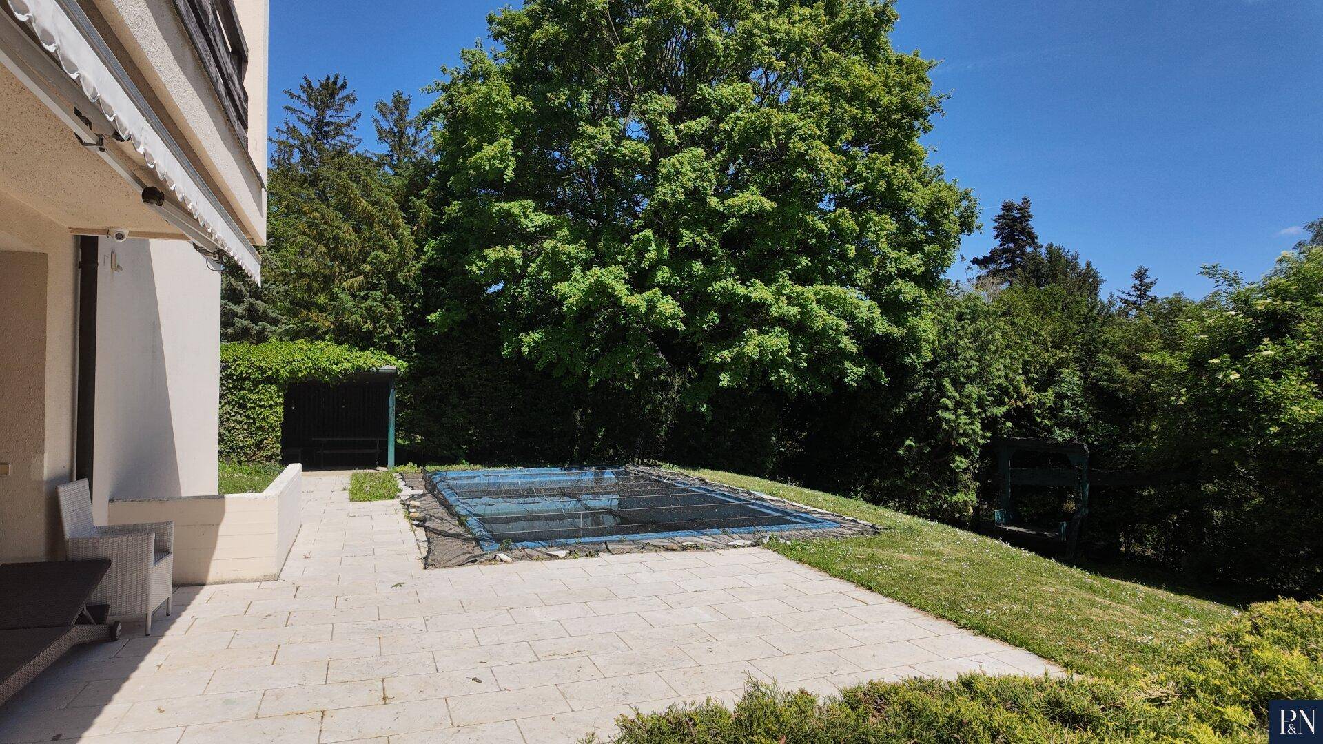 Gepflasterte Terrasse neben dem Haus, die zum Garten mit dem abgedeckten Pool führt.