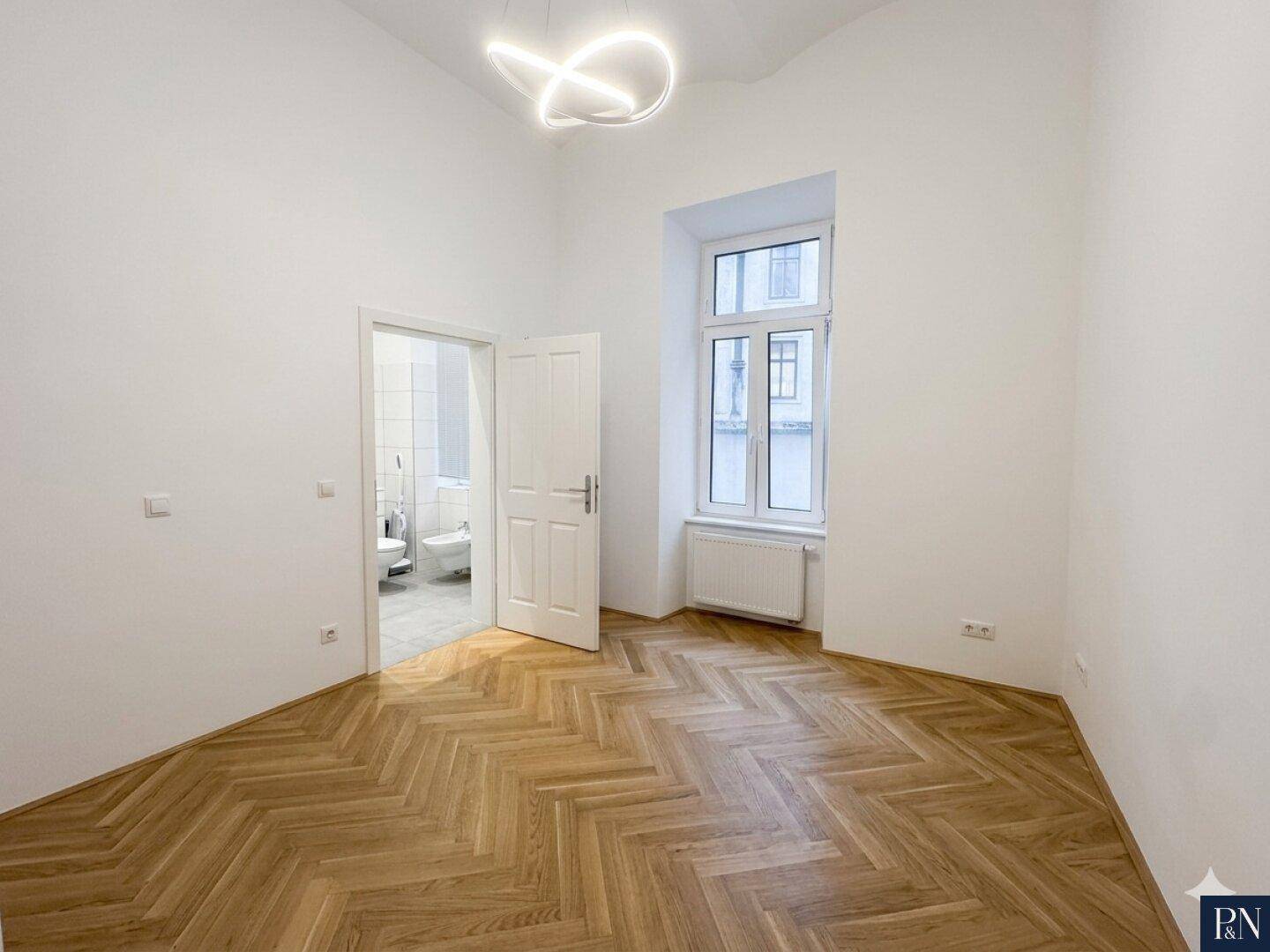 Helles Zimmer mit Fischgrätparkett und Fenster, bietet Zugang zu einem modernen Badezimmer.