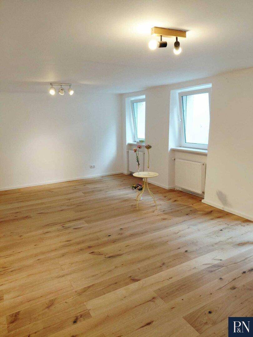 Geräumiger, heller Raum mit Holzboden und zwei Fenstern, ausgestattet mit Deckenleuchten.