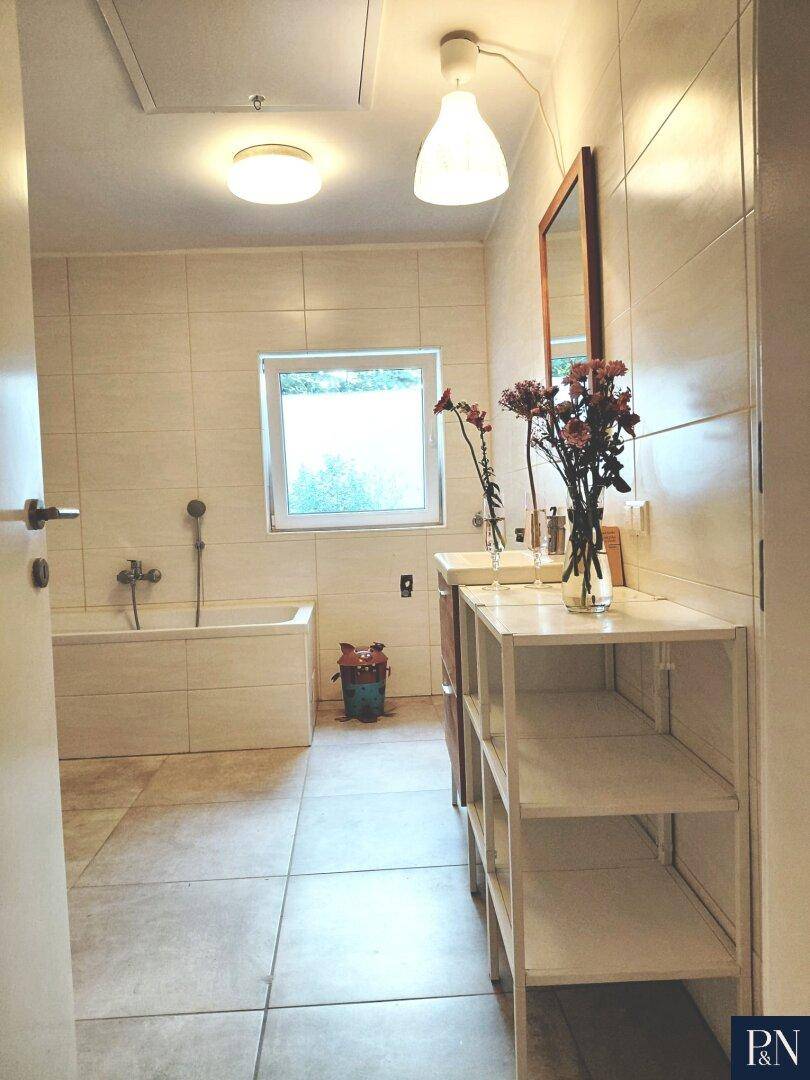 Modernes Badezimmer mit Badewanne, Fenster und hellen Fliesen, ausgestattet mit einem Spiegel.