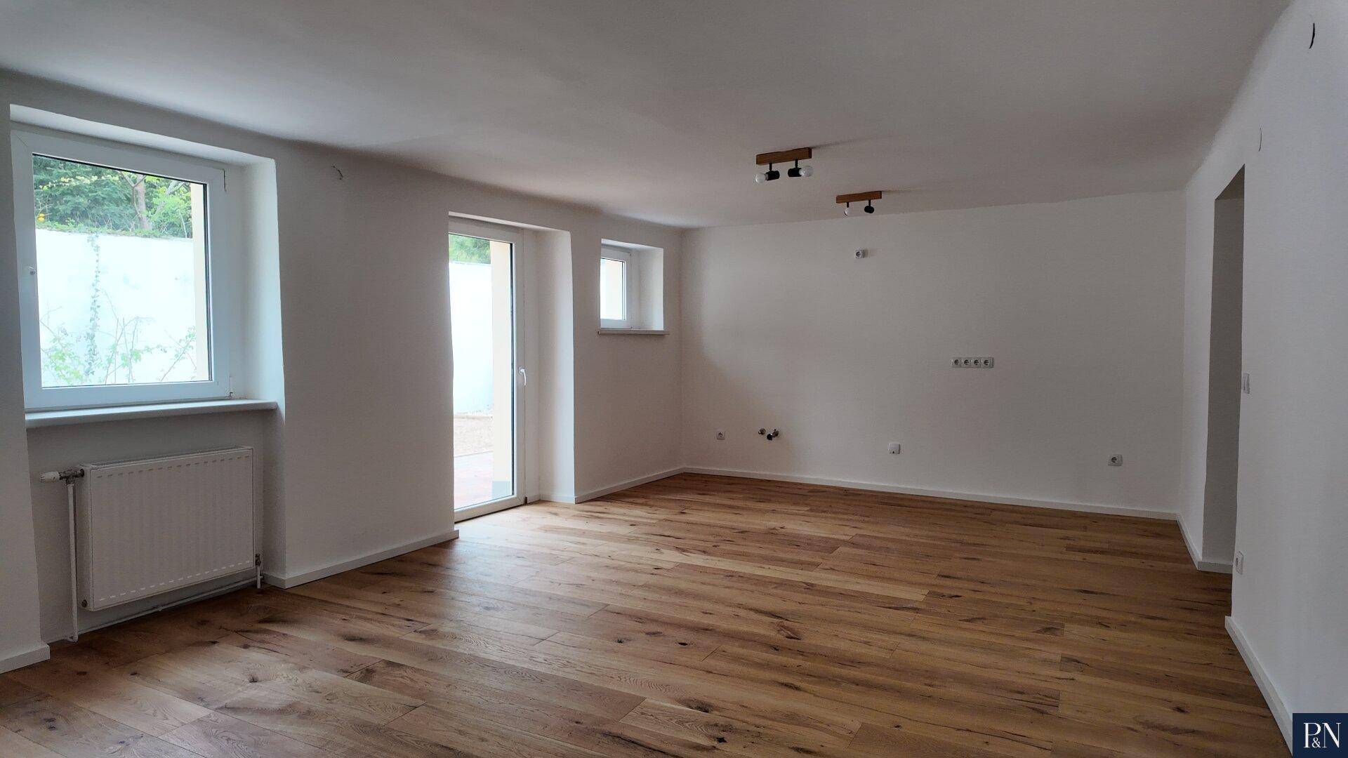 Geräumiger Wohnbereich mit Holzboden, großen Fenstern und Zugang zu einem Außenbereich.