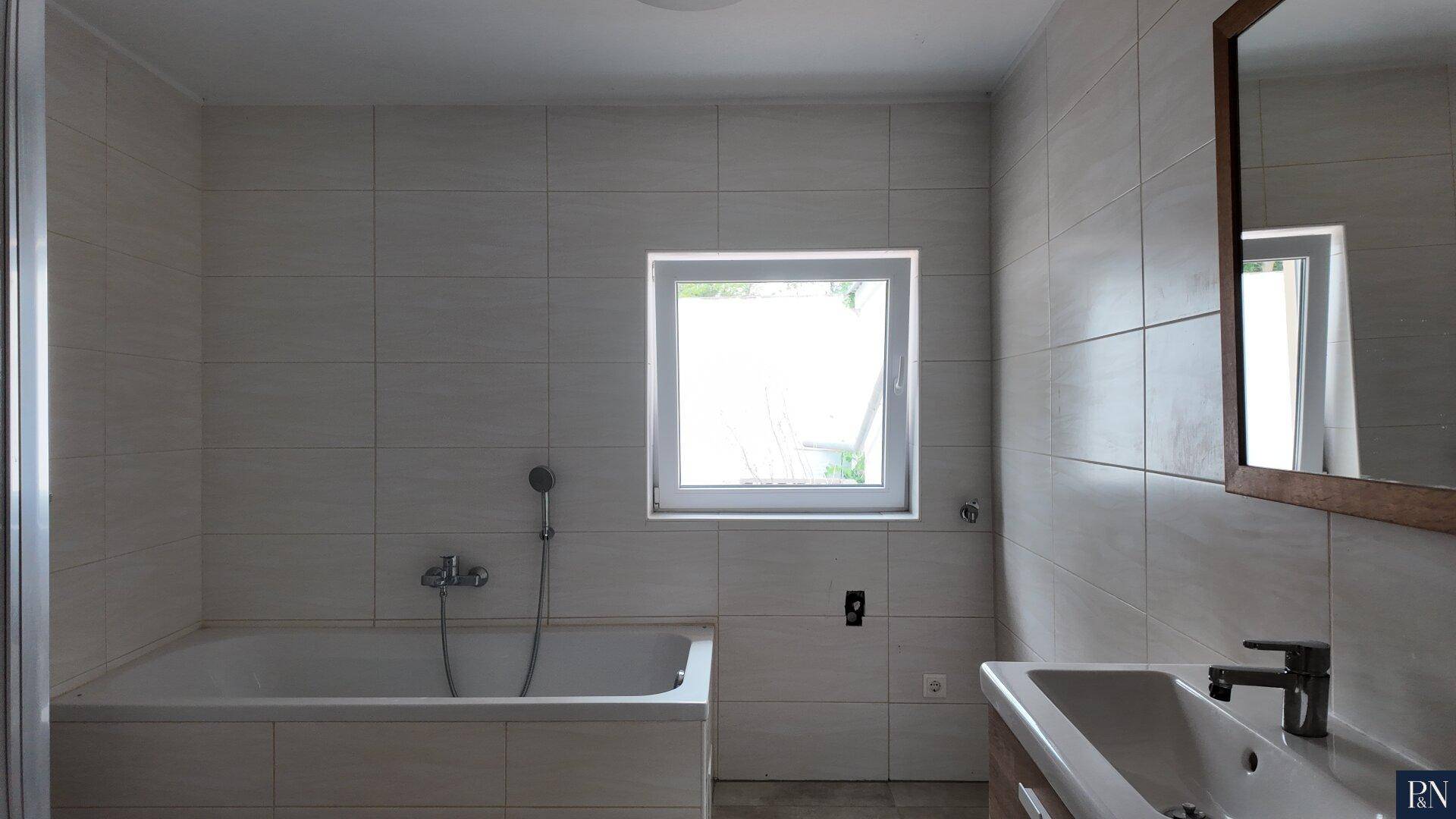 Helles Badezimmer mit Badewanne, Fenster und einem Waschbecken, umgeben von hellen Fliesen.