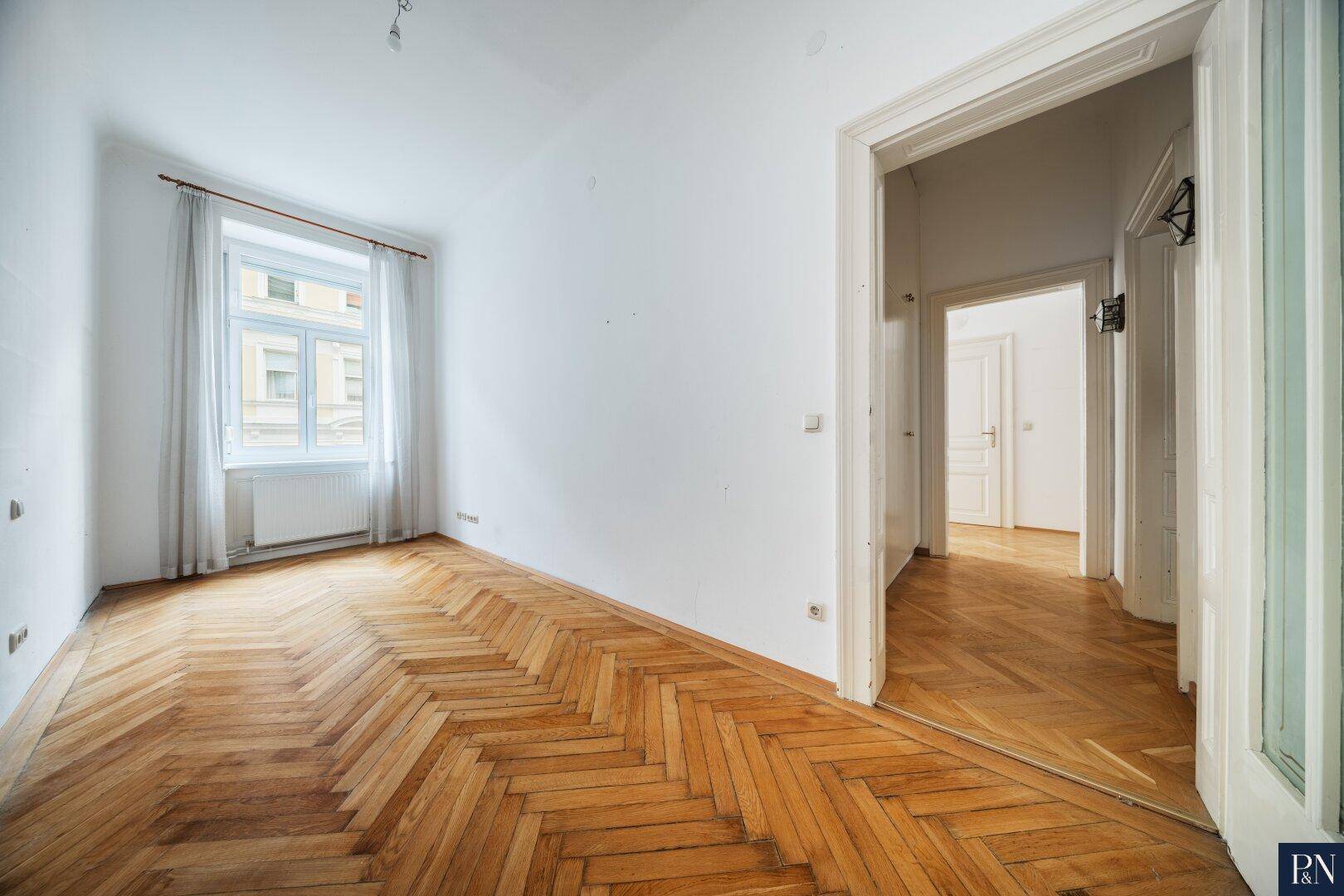 Helles Zimmer mit Fischgrätparkett, hohen Decken und einem Fenster mit Blick auf die Stadt.