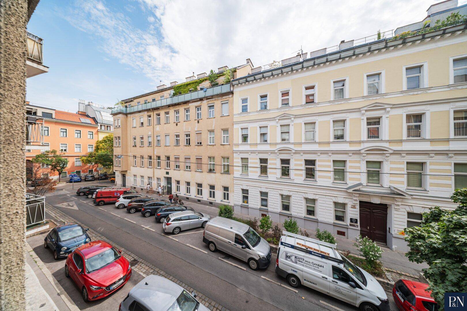 Blick von einem Balkon auf eine belebte Stadtstraße mit parkenden Autos und historischen Gebäuden.