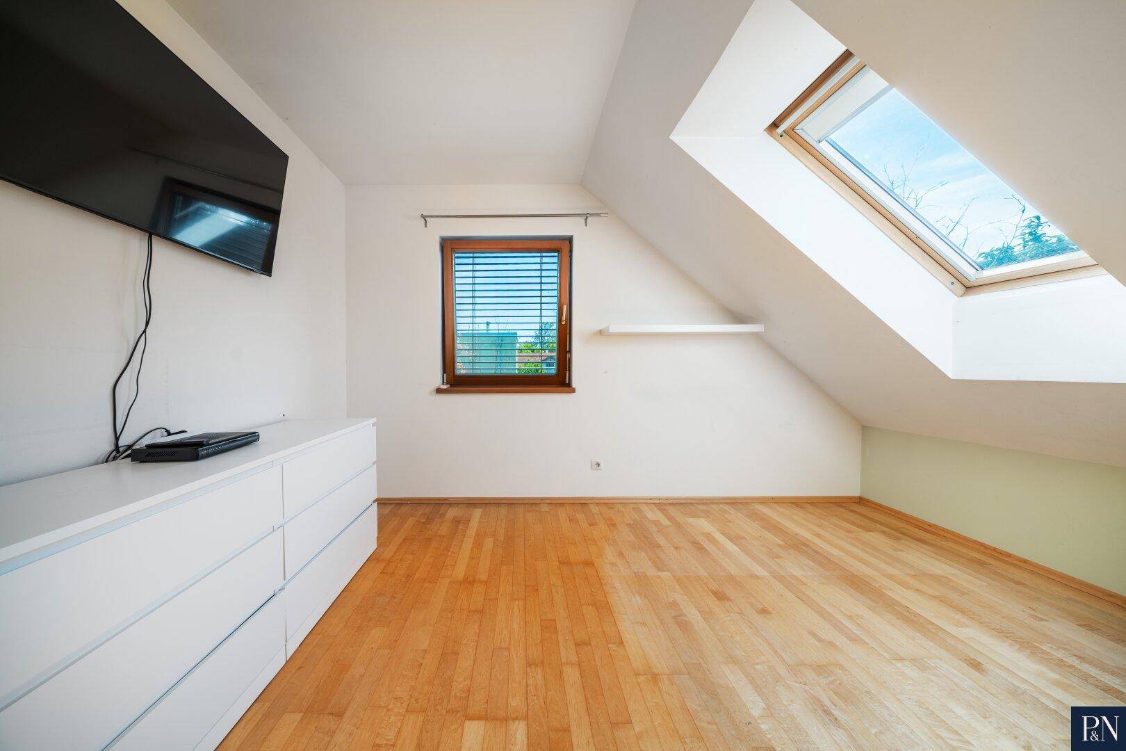 Helles Schlafzimmer im Dachgeschoss mit Parkettboden, Dachfenster und TV-Wandhalterung.