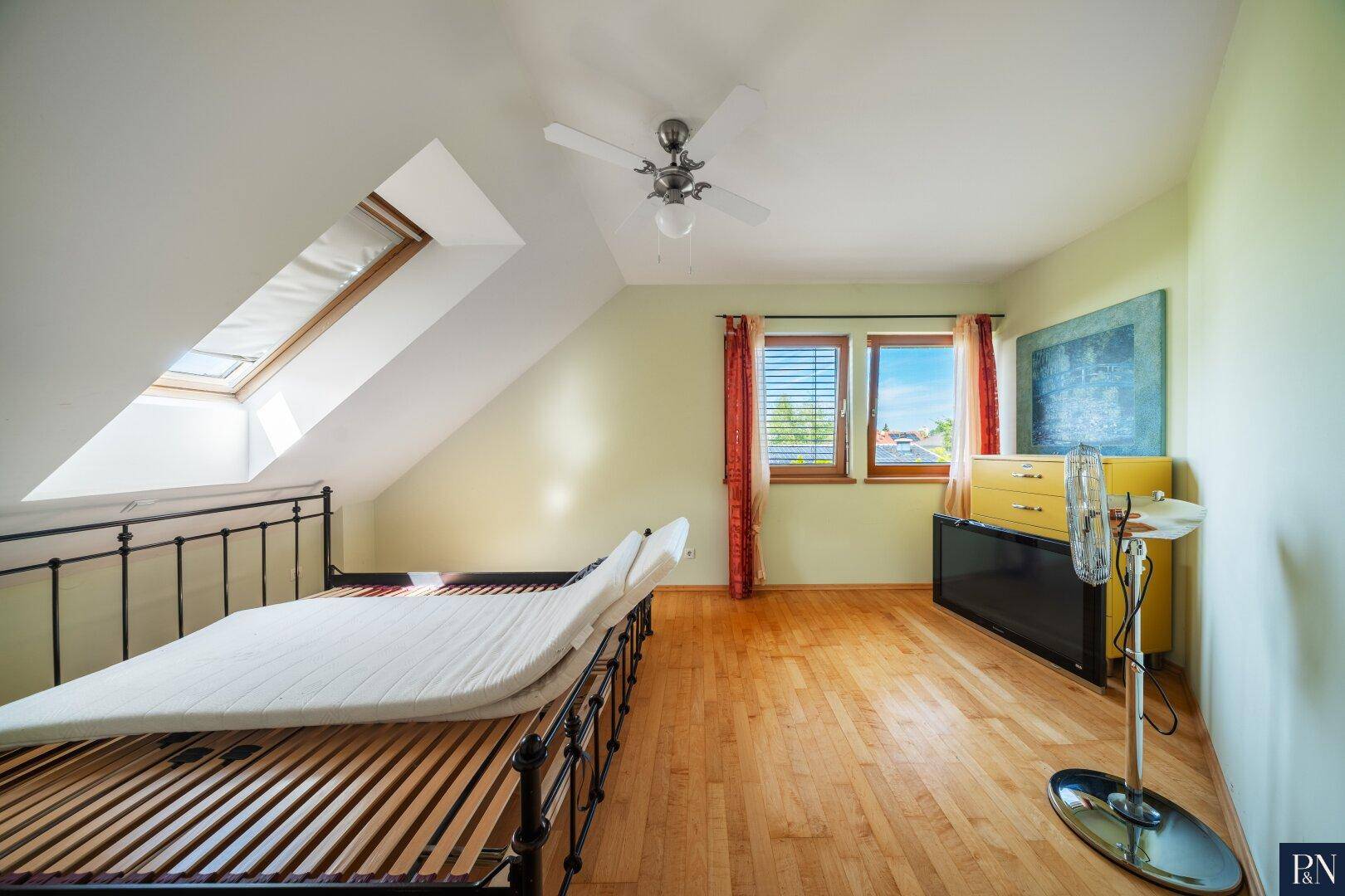Schlafzimmer mit Holzboden, Dachschräge, Deckenventilator und zwei Fenstern.