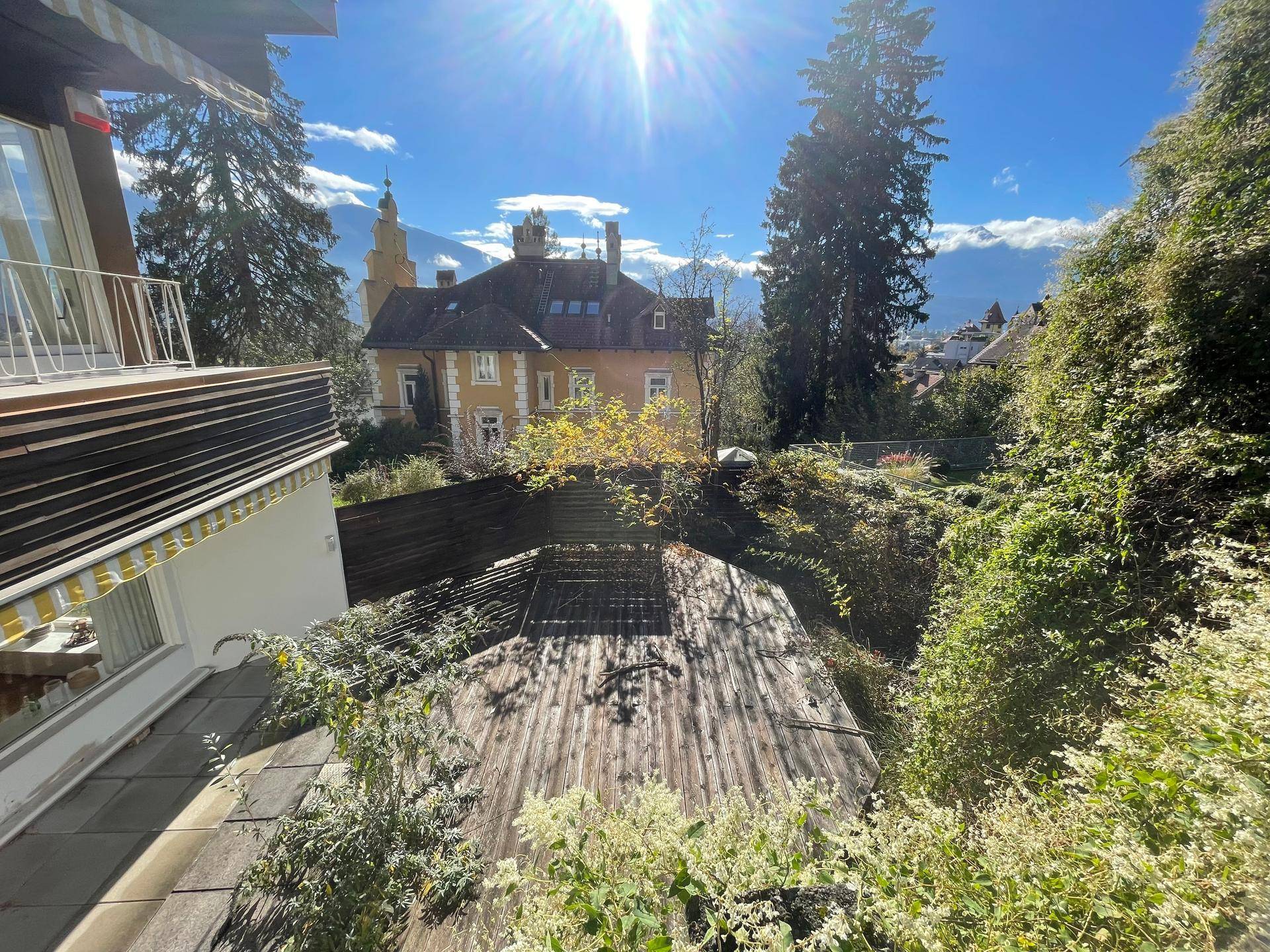 Aussicht von einem Balkon auf ein Nachbargebäude, einen Garten und die umliegende Berglandschaft.