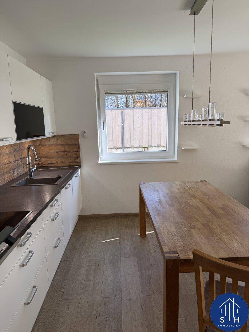 Helle Küche mit weißen Schränken, Holzarbeitsplatte, Spüle und einem Esstisch unter dem Fenster.
